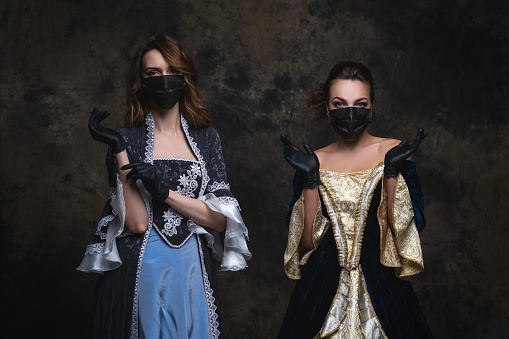 Two women in renaissance dress, face mask and gloves on abstract dark background, coronavirus, covid-19 protection concept, studio shot