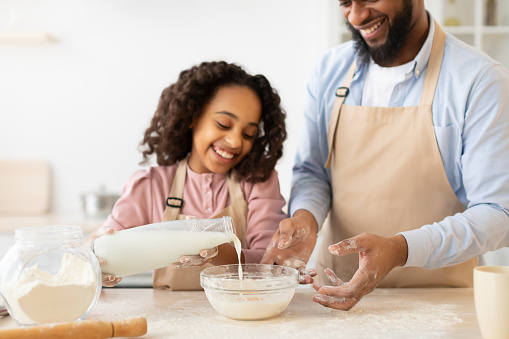 Cheerful Black Family Father And Daughter Baking Together In Kitchen. Cute Little African American Girl Adding Ingregients To Bowl, Pouring Milk, Preparing Dough For Cookies With Daddy At Home