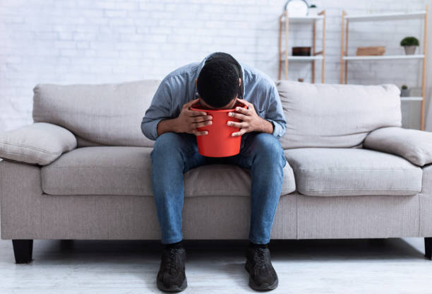 uomo africano che vomita nel secchio seduto sul divano a casa - abdomen addiction adult alcohol foto e immagini stock
