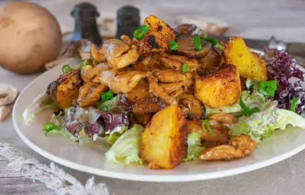 Delicious and tasty homemade chicken dish with fried and chopped hen breast served with roasted potatoes, mixed salad and a creamy mushroom sauce on a plate on kitchen table background