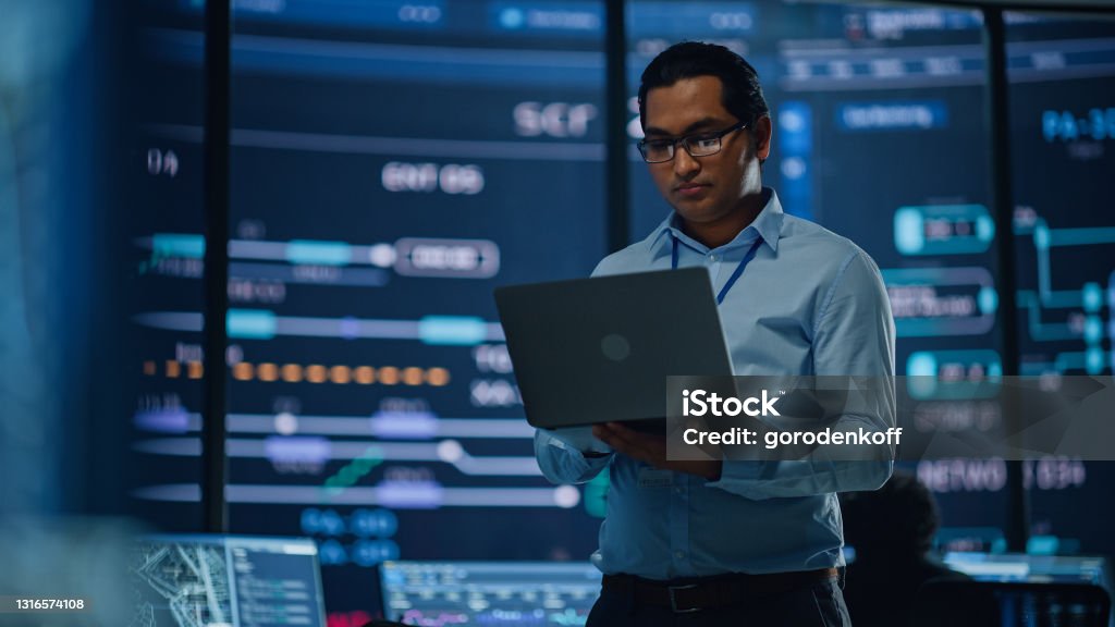Young Multiethnic Male Government Employee Uses Laptop Computer in System Control Monitoring Center. In the Background His Coworkers at Their Workspaces with Many Displays Showing Technical Data. Technology Stock Photo
