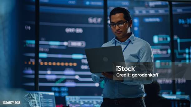 Young Multiethnic Male Government Employee Uses Laptop Computer In System Control Monitoring Center In The Background His Coworkers At Their Workspaces With Many Displays Showing Technical Data Stock Photo - Download Image Now