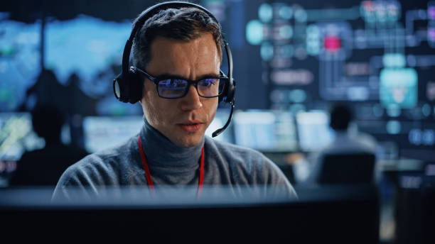 istock Portrait of Professional IT Technical Support Specialist Working on Computer in Monitoring Control Room with Digital Screens. Employee Wears Headphones with Mic and Talking on a Call. 1316574129