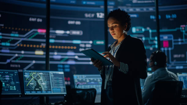 istock Young Multiethnic Female Government Employee Uses Tablet Computer in System Control Monitoring Center. In the Background Her Coworkers at Their Workspaces with Many Displays Showing Technical Data. 1316574130