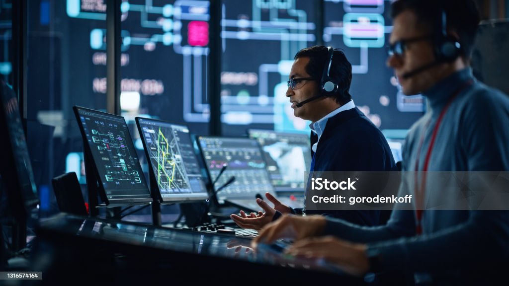 Portrait of Professional IT Technical Support Specialist Working on Computer in Monitoring Control Room with Digital Screens. Employee Wears Headphones with Mic and Talking on a Call. Network Security Stock Photo