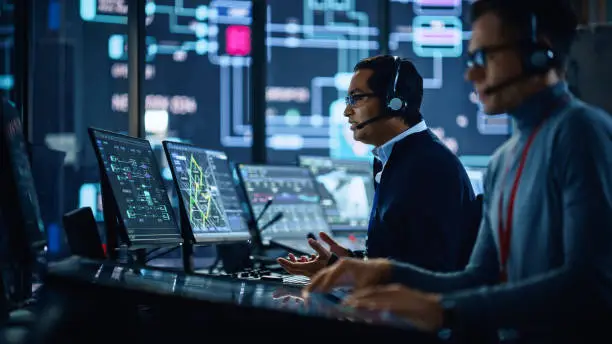 Photo of Portrait of Professional IT Technical Support Specialist Working on Computer in Monitoring Control Room with Digital Screens. Employee Wears Headphones with Mic and Talking on a Call.