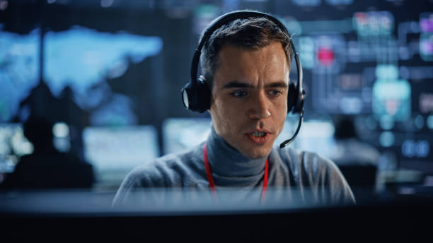 istock Portrait of Professional IT Technical Support Specialist Working on Computer in Monitoring Control Room with Digital Screens. Employee Wears Headphones with Mic and Talking on a Call. 1316574172