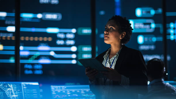 istock Young Multiethnic Female Government Employee Uses Tablet Computer in System Control Monitoring Center. In the Background Her Coworkers at Their Workspaces with Many Displays Showing Technical Data. 1316574200