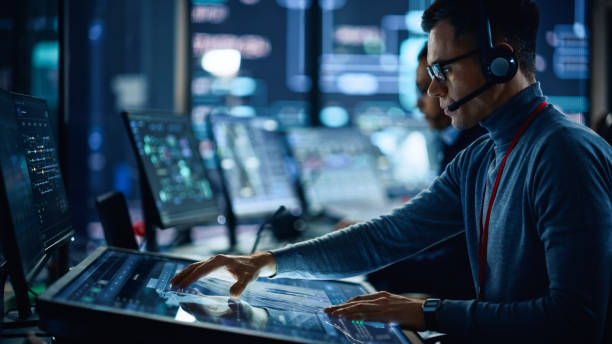 istock Portrait of Professional IT Technical Support Specialist Working on Computer in Monitoring Control Room with Digital Screens. Employee Wears Headphones with Mic and Talking on a Call. 1316574222
