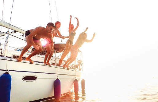 Side view of young crazy friends jumping from sailboat on sea ocean trip - Men and women having summer fun together at sail boat party day - Luxury excursion concept on warm backlight filter