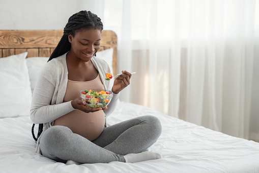 Pregnant Woman Nutrition. Happy Black Expecting Lady Eacting Fresh Vegetable Salad While Sitting In Bed, Smiling African American Female Enjoying Healthy Organic Food During Pregnancy, Copy Space