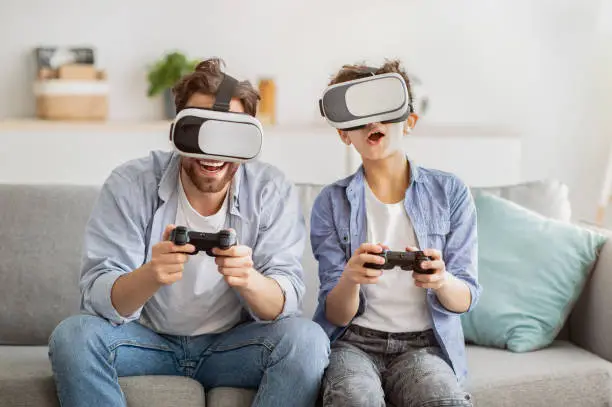 Virtual reality. Excited father and teen son trying VR glasses and playing videogames together, holding joysticks, sittin gon sofa at home interior. Leisure time with dad
