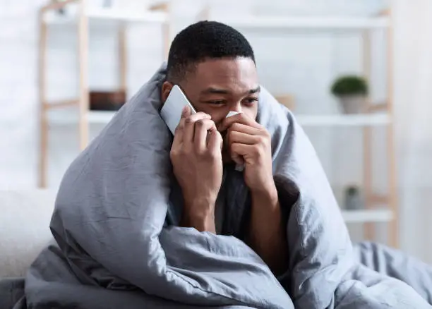 Photo of Ill Black Man Calling Doctor Having Runny Nose Sitting Indoors