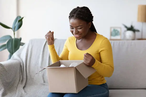 Happy black woman unboxing cardboard box parcel at home, emotionally reacting to successful shopping, satisfied with great purchase, clenching fists with excitement, sitting on couch in living room