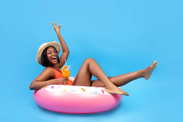 Positive black woman sitting on inflatable ring, holding tropical cocktail on blue background, full length portrait Positive black woman sitting on inflatable ring, holding tropical cocktail on blue background, full length. Happy African American lady having beach party with pool float and refreshing summer drink black women in bathing suits stock pictures, royalty-free photos & images