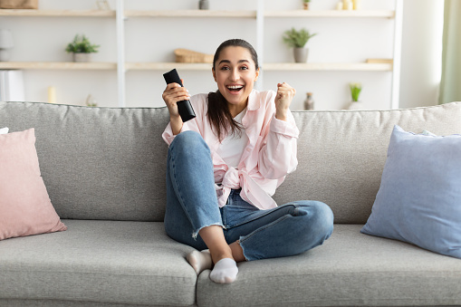 Yes. Excited young woman watching TV show or movie, holding remote contoller, sitting on the couch at home in living room. Happy lady cheering, shaking clenched fists, making winner gesture