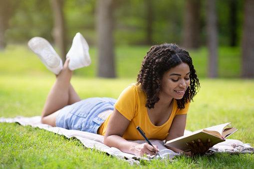 African American female student writing down notes from book while lying on blanket at park