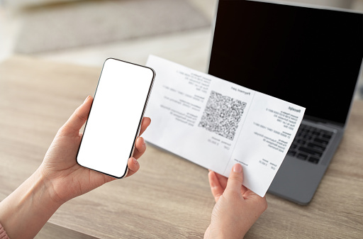 Female hands holding smartphone and scanning the QR code from paper bill, cellphone with white blank screen. Paying online from home, modern technologies concept