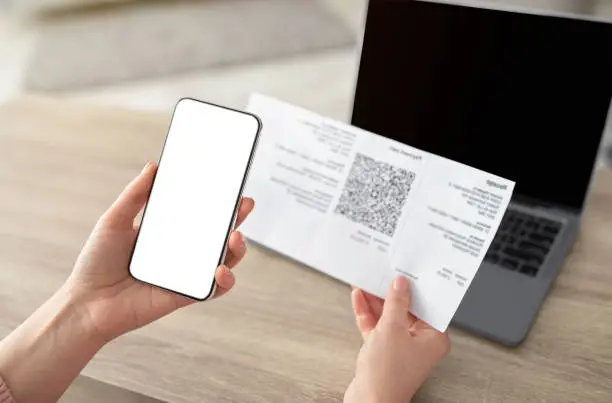 Female hands holding smartphone and scanning the QR code from paper bill, cellphone with white blank screen. Paying online from home, modern technologies concept