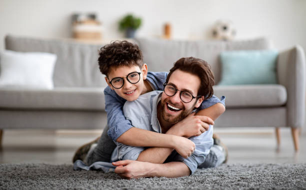 Parents and children being friends. Joyful father and son having fun, dad lying on floor, carrying boy on back Parents and children being friends. Joyful father and son having fun, dad lying on floor, carrying boy on back and smiling together to camera, spending tim at home together. Single dad concept eyeglasses stock pictures, royalty-free photos & images