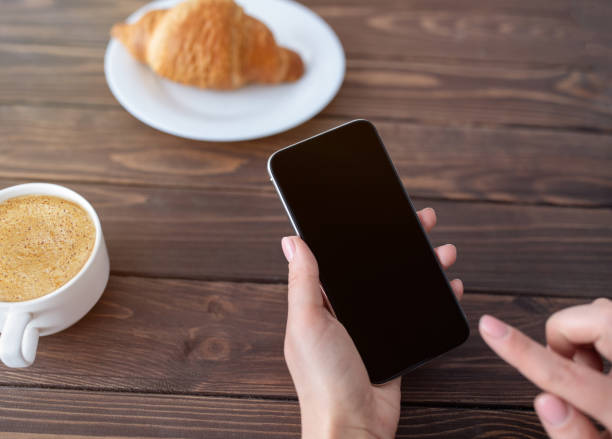 istock Woman using smartphone with black blank screen while drinking coffee and eating croissant at cafe, mockup for design 1316585422