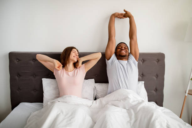 joven pareja multirracial que se estira en la cama después de despertarse en casa - stretching boyfriend indoors lifestyles fotografías e imágenes de stock