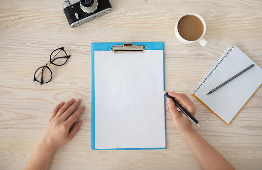 Female hands writing plan for trip in empty notepad at the wooden desk, top view, copy space for text. Eyeglasses, retro photocamera, cup of coffee and spiral notebook on table, flat lay