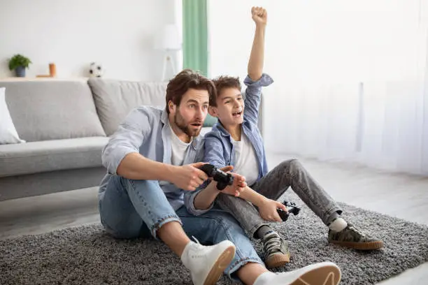 Father-son competition. Joyful boy winning dad in videogame and raising hand, celebrating victory. Family having fun at home, sitting on floor carpet, empty space