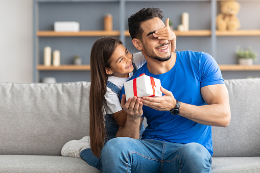 This Present Is For You. Portrait of loving girl with long hair holding and giving wrapped gift box, making surprise and covering dad's eyes, greeting with father's day or birthday, sitting on couch