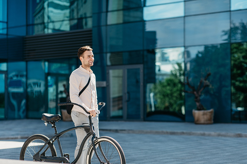 Office worker, active and healthy lifestyle in urban. Cheerful millennial handsome male manager in stylish clothes walks with bicycle near modern office building, outdoor, side view, free space