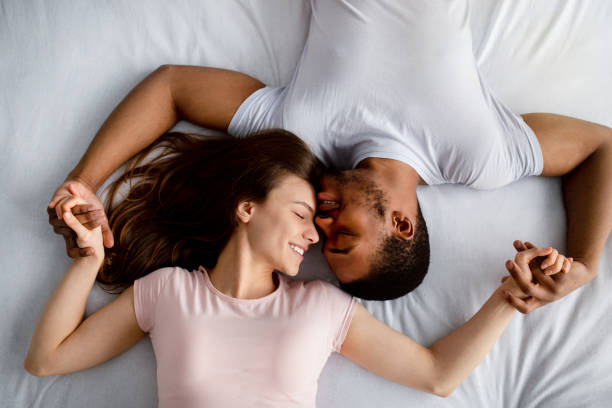 вид сверху на молодую многорасовую пару, лежащую на кровати, обнимающаяся, выражаюющая привязанность друг к другу - sexual activity black couple african descent стоковые фото и изображения