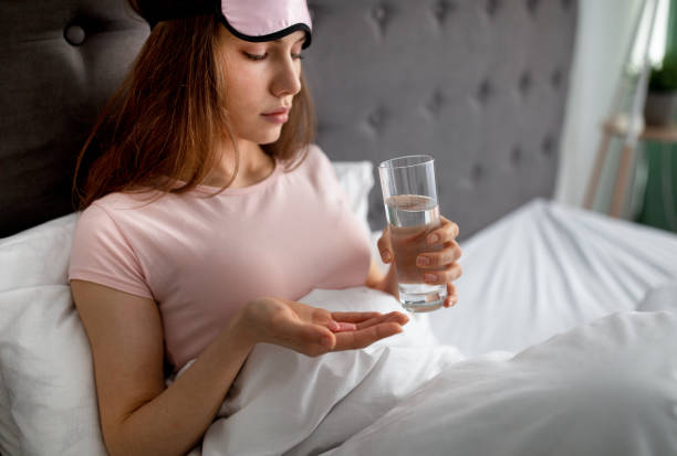 Upset young woman sitting in bed with glass of water, taking sleeping pills or painkillers at home, copy space Upset young woman sitting in bed with glass of water, taking sleeping pills or painkillers at home, copy space. Millennial lady drinking medications, not feeling well indoors sleeping pill stock pictures, royalty-free photos & images