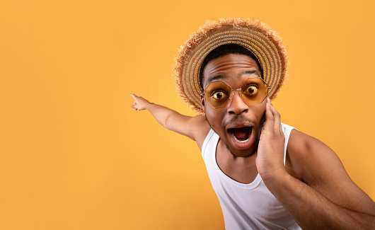 Summer sale. Shocked black guy in straw hat pointing at empty space on yelow studio background. Surprised African American man advertising your summertime product or service