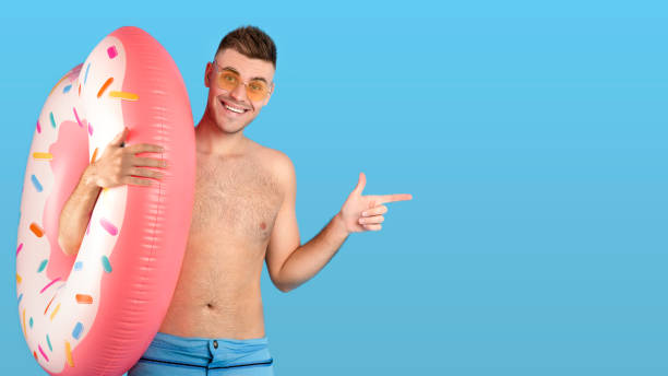 istock Smiling young guy in shorts with inflatable ring pointing aside at empty space over blue background, banner design 1316589002