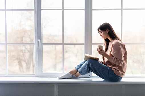 Free Time To Relax. Profile of beautiful woman sitting on white windowsill at home, reading paper book, holding cup and drinking hot coffee. Happy lady taking break with mug near window, copy space