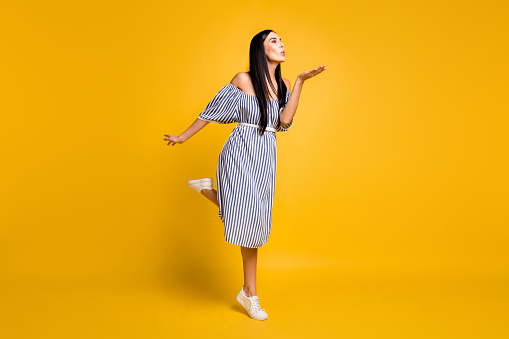 Full length photo of charming sweet young lady wear striped dress sending kiss arm empty space isolated yellow color background.