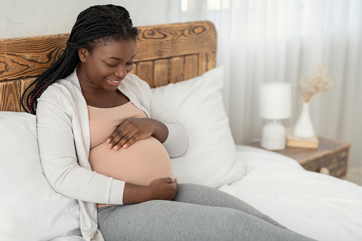 Happy Expectation. Portrait Of Young Pregnant African American Lady Relaxing At Home, Positive Black Woman Sitting On Bed Touching Her Belly And Smiling,