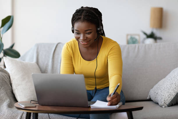 Smiling black lady in headset writing notes while watching webinar on laptop Smiling black lady in headset writing notes while watching webinar on laptop at home, young african female student in headphones enjoying distance learning, having online lesson with teacher mba online stock pictures, royalty-free photos & images