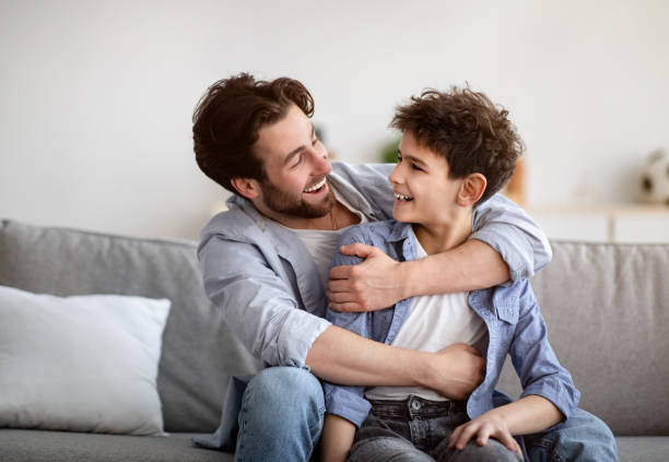 istock Two generations concept. Happy father embracing son, looking at each other and smiling, sitting on sofa at home 1316604468