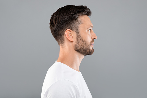 Profile portrait of middle-aged bearded man in white t-shirt over grey studio background, copy space. Side view of handsome confident man posing on gray, standing straight and looking aside