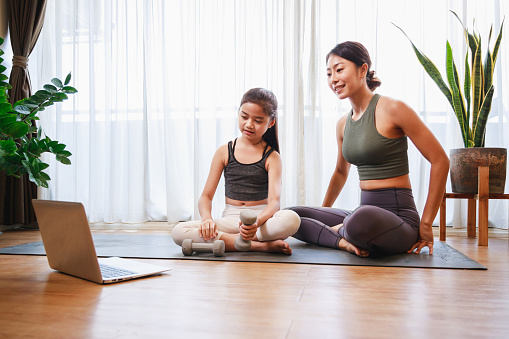 Mom and Little Girl doing dumbbell exercises by using online learning with a laptop computer on a yoga mat in the living room at home. New normal concept. Healthy lifestyle. Relaxing at home