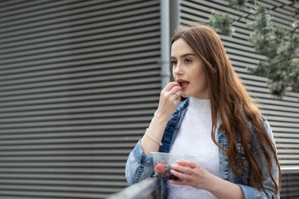 istock Young woman eating fruit from a plastic (polyethylene terephthalate) container, standing by a metal-clad building - she is wearing a floral skirt, white jumper and a denim jacket 1316619264