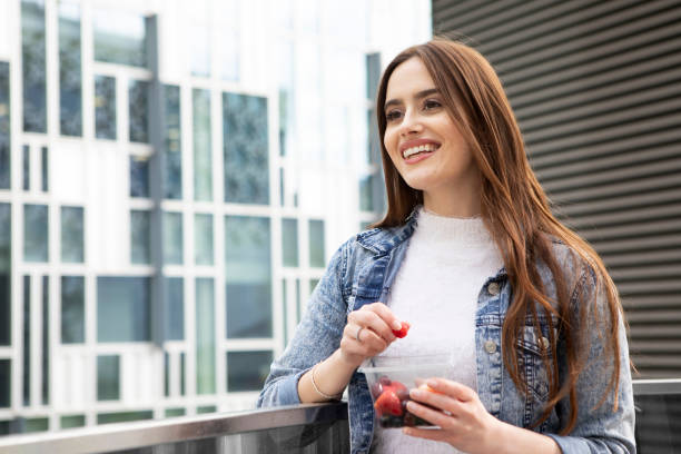 istock Young woman eating fruit from a plastic (polyethylene terephthalate) container, standing with office buildings behind, and smiling - she is wearing a floral skirt, white jumper and a denim jacket 1316619285