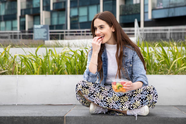 istock Young woman eating fruit from a plastic (polyethylene terephthalate) container, sitting cross-legged on a concrete seat, with plants behind - she is wearing a floral skirt, white jumper and a denim jacket 1316619380