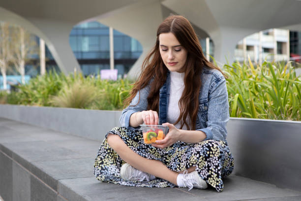 istock Young woman eating fruit from a plastic (polyethylene terephthalate) container, sitting cross-legged on a concrete seat, with plants behind - she is wearing a floral skirt, white jumper and a denim jacket 1316619397