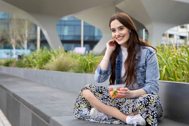 istock Young woman eating fruit from a plastic (polyethylene terephthalate) container, sitting cross-legged on a concrete seat, with plants behind - she is wearing a floral skirt, white jumper and a denim jacket 1316619420