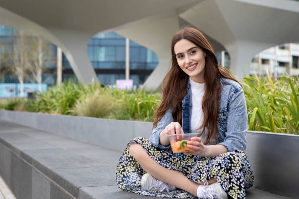 istock Young woman eating fruit from a plastic (polyethylene terephthalate) container, sitting cross-legged on a concrete seat, with plants behind - she is wearing a floral skirt, white jumper and a denim jacket 1316619421