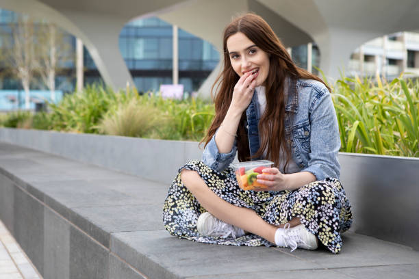 istock Young woman eating fruit from a plastic (polyethylene terephthalate) container, sitting cross-legged on a concrete seat, with plants behind - she is wearing a floral skirt, white jumper and a denim jacket 1316619425