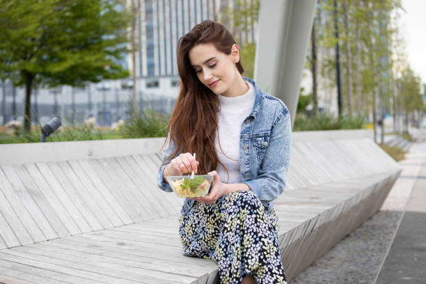 Young woman eating a chicken and pasta salad from a plastic (polyethylene terephthalate) container, sitting on a modern public bench, with city buildings behind - she is wearing a floral skirt, white jumper and a denim jacket