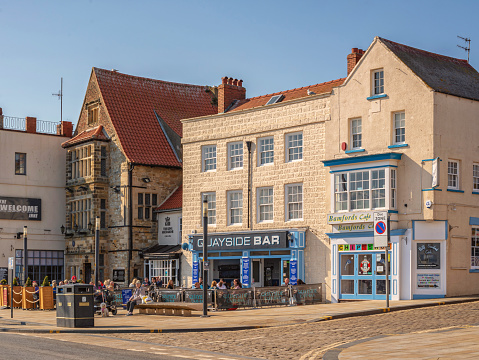 Scarborough, UK.  April 30, 2021. A road with bars and cafes with differing styles of architecture.  People sit outside in the sunshine and a clear sky is overhead.